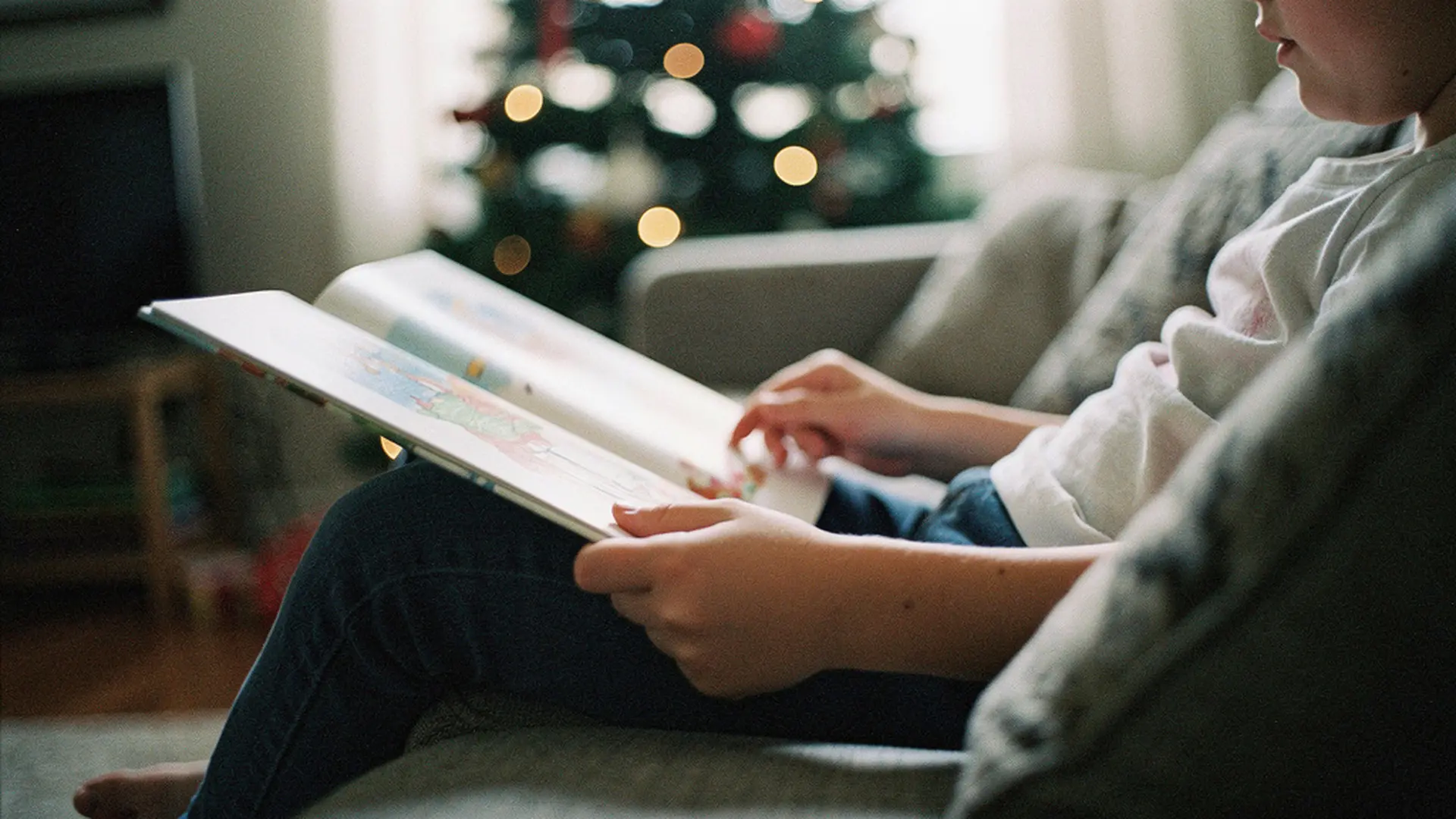 Enfant tenant un livre illustré ouvert sur ses genoux près du sapin de Noël, gros plan sur les mains