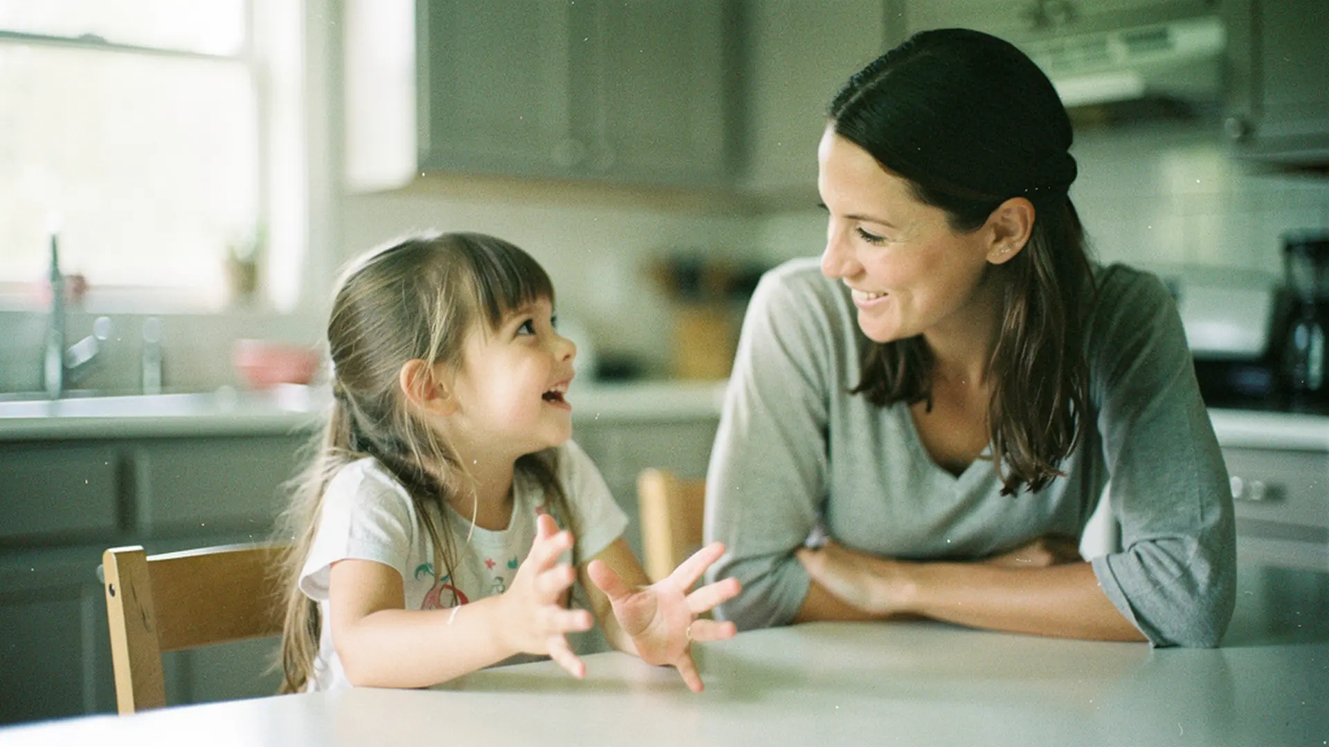 Mère et sa fille de 4 ans attablées dans une cuisine, la fillette parlant avec animation pendant que sa mère l'écoute