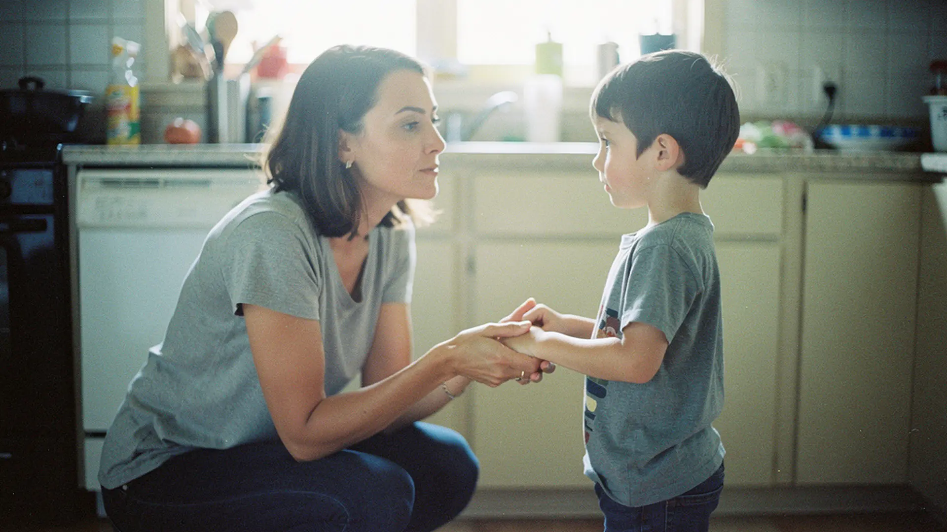 Mère accroupie face à son fils de 5 ans, lui tenant les mains, expression calme et attentive dans une cuisine