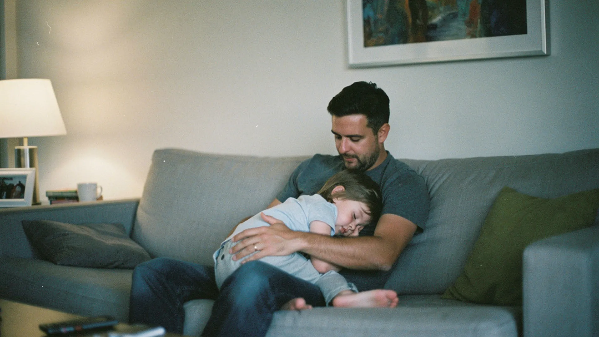 Père assis sur un canapé le soir, enfant de 3 ans endormi contre lui, lumière douce, expression fatiguée mais apaisée