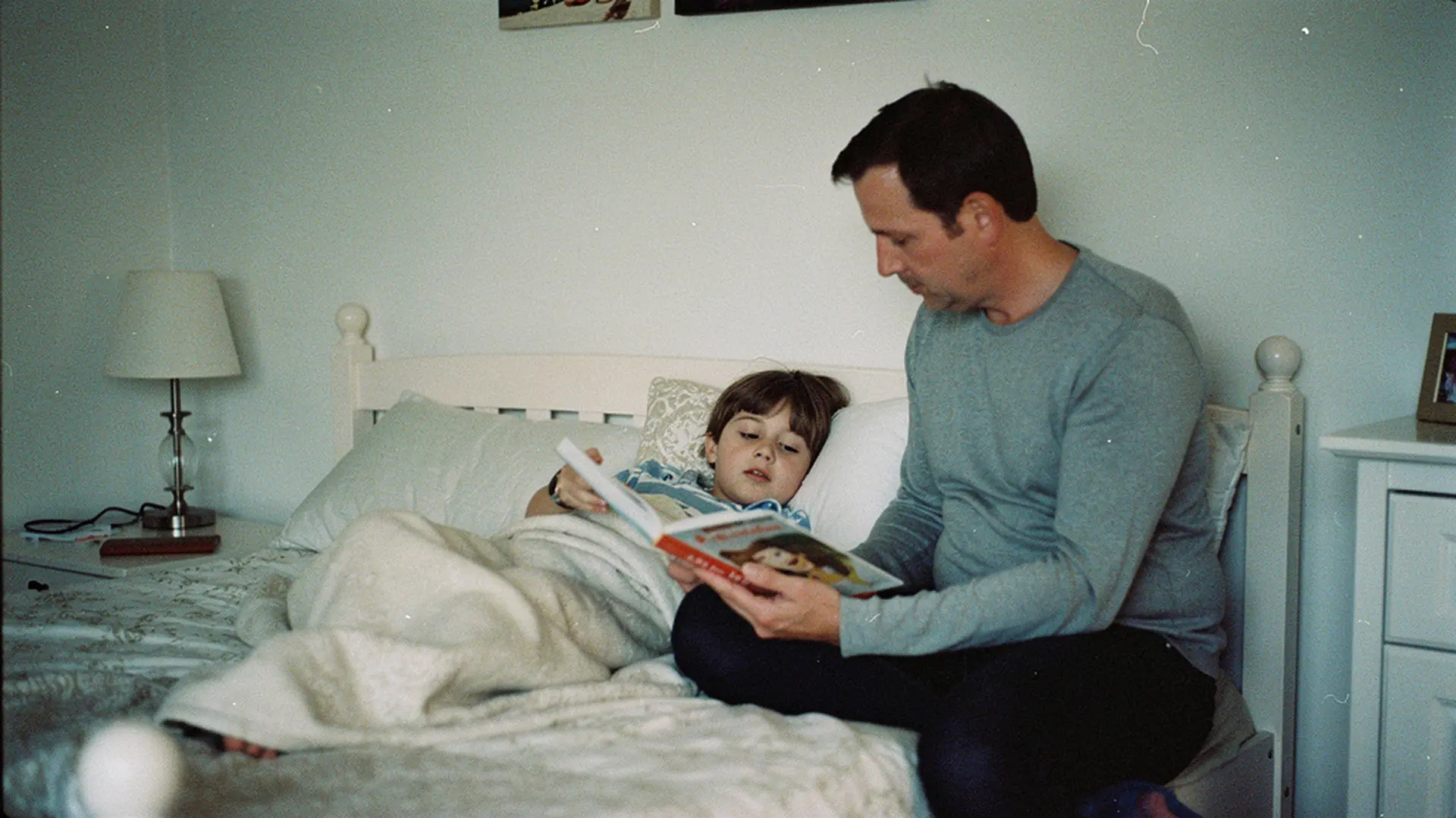Parent assis au bord du lit lisant une histoire à son enfant sous la couette, lumière tamisée de la chambre